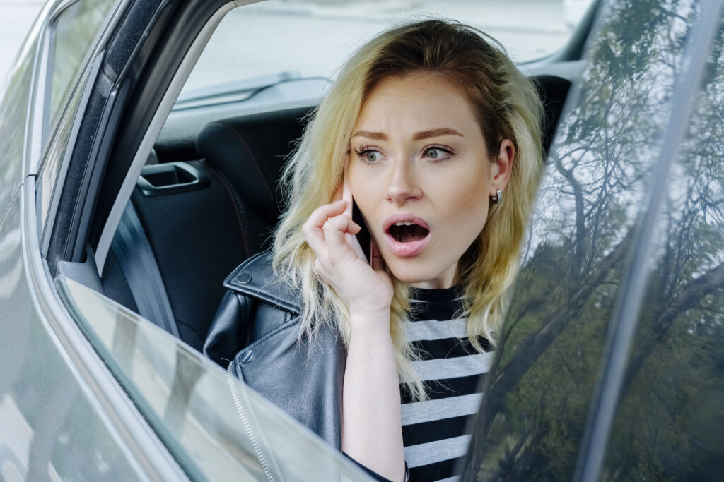 young surprised woman talking on smartphone while travelling in the back seat of car wearing striped top and black leather jacket