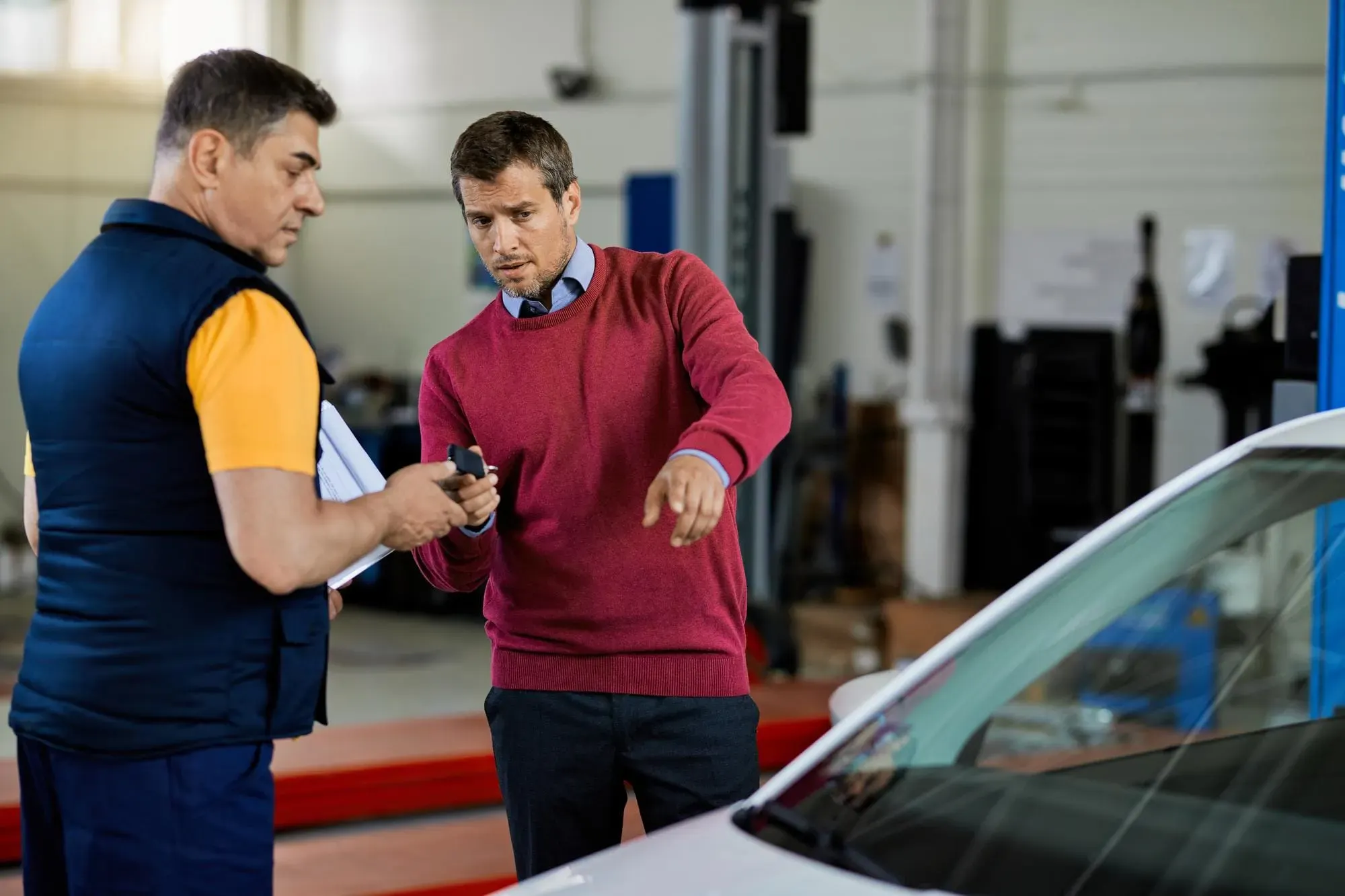 Customer speaks with an auto technician in a repair shop while pointing toward a car during a vehicle inspection.