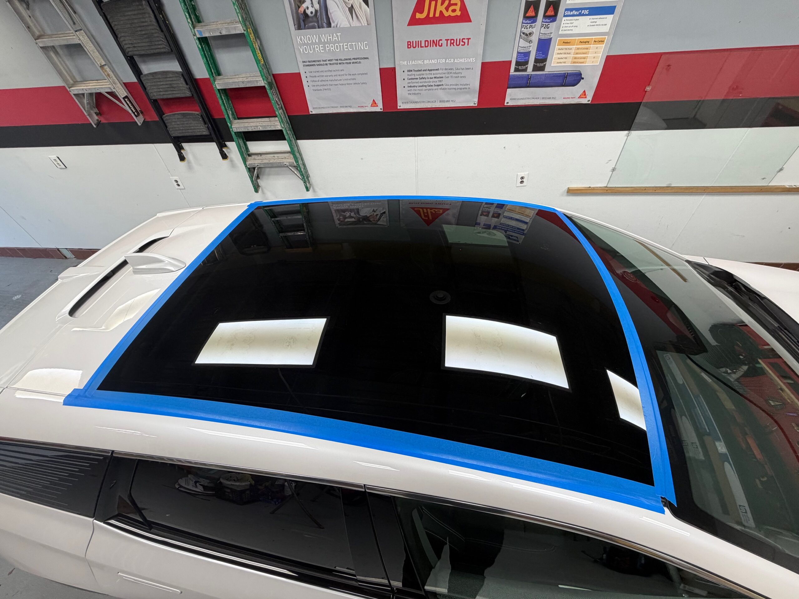 Top view of a white vehicle with a panoramic sunroof secured using blue protective tape during a professional auto glass installation in a repair shop