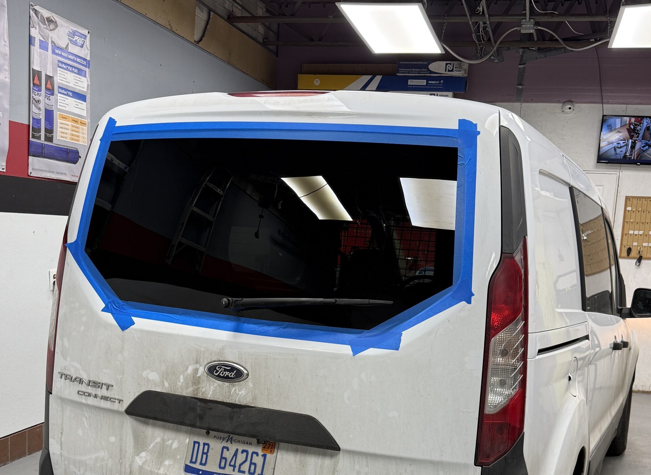 Rear view of a white Ford Transit Connect van with a newly installed back glass and blue tape around the edges, at the Save On Auto Glass shop
