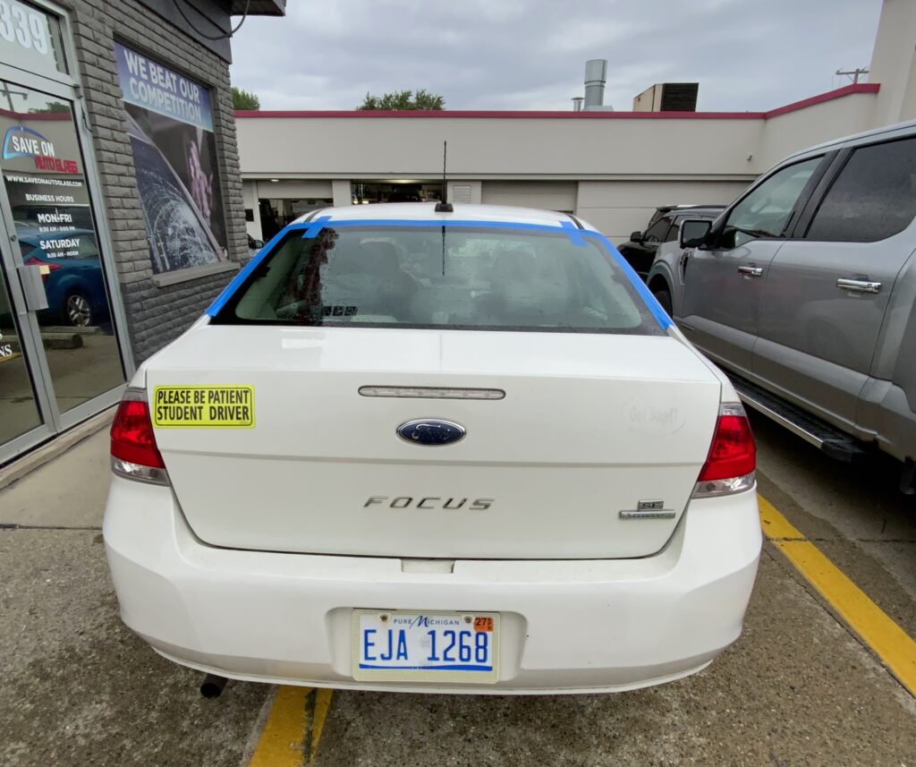 White Ford Focus sedan with back glass replaced by Save On Auto Glass, blue tape still around the edges of the new glass, parked outside the shop