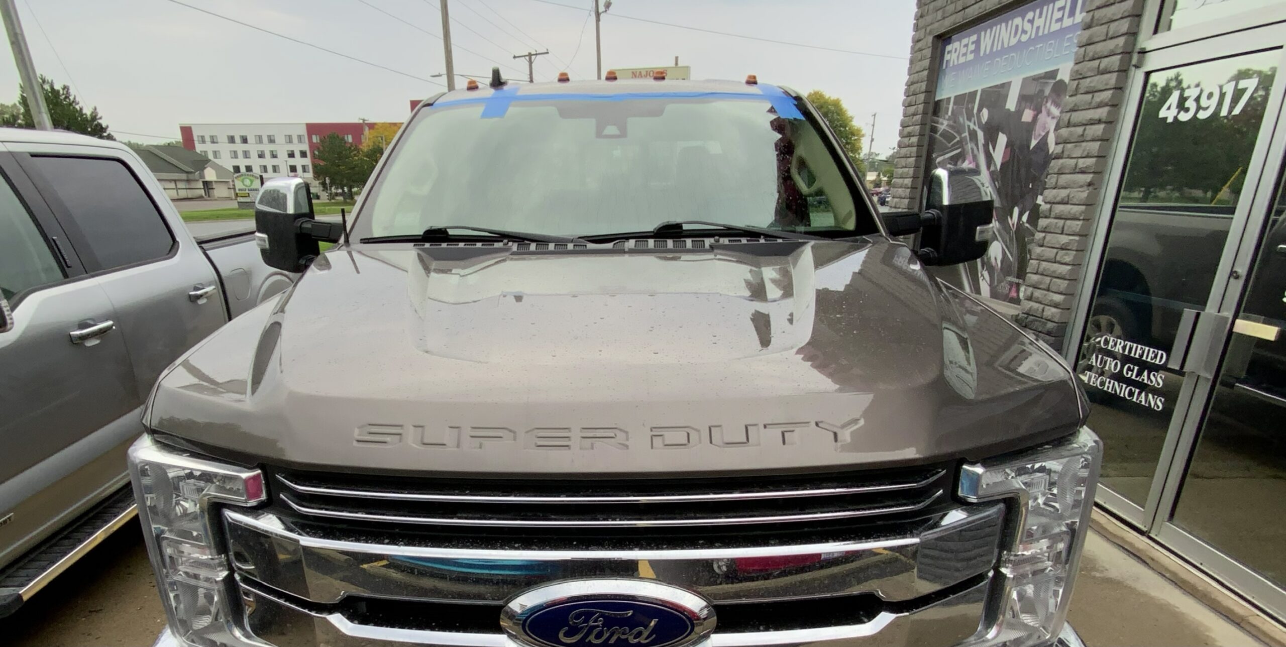 Gray Ford vehicle with windshield replaced by Save On Auto Glass, blue tape still along the top edge of the new glass, parked in front of the shop
