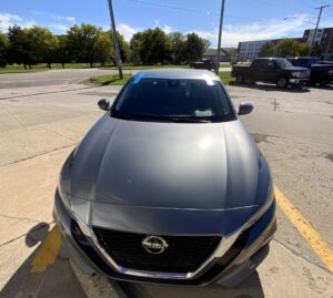 Black car parked outdoors in a parking lot in Fenton, Michigan, showing the windshield and exterior exposed to direct sunlight and outdoor weather conditions.