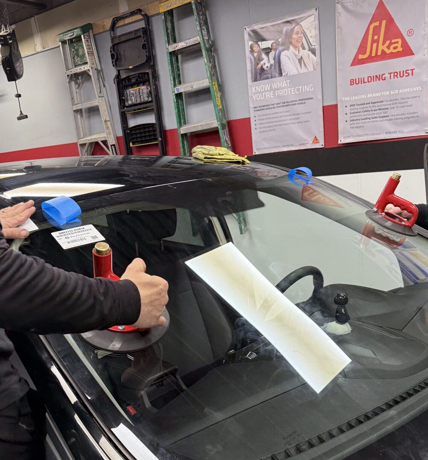 Technician using red suction cup handles to position a new windshield on a black car inside the Save On Auto Glass shop