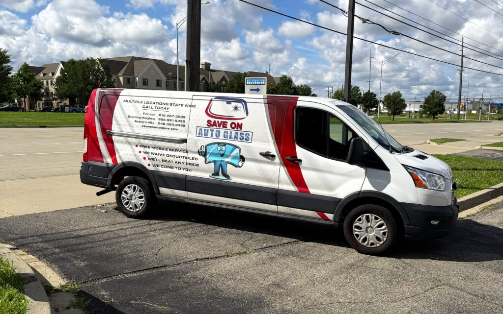 Save On Auto Glass white mobile service van with red accents parked in a parking lot on a sunny day under a blue sky with clouds