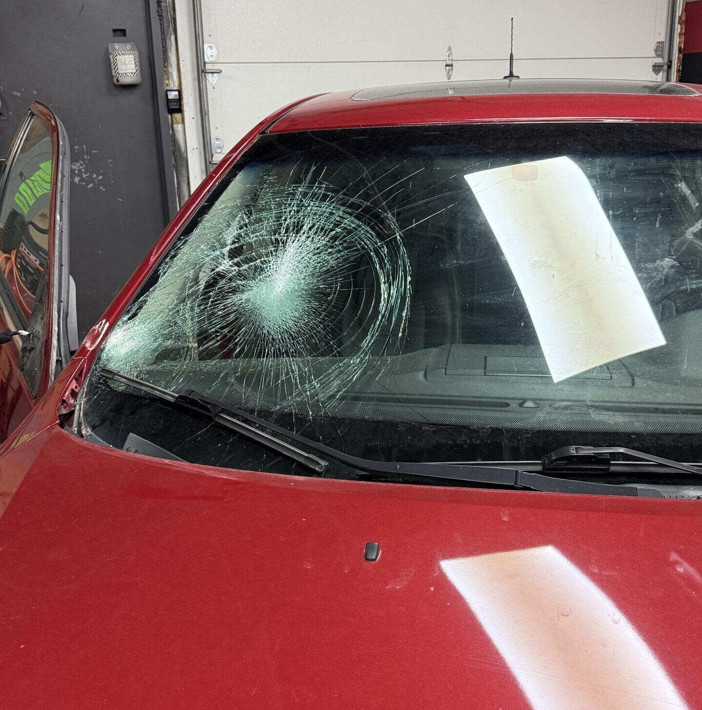 Red car with a badly cracked windshield showing a large spiderweb impact, waiting for replacement